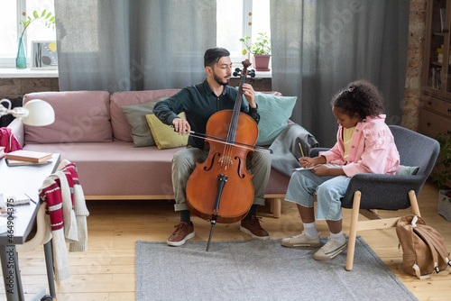 Young teacher of music showing how to play cello to biracial pupil sitting in armchair in front of him