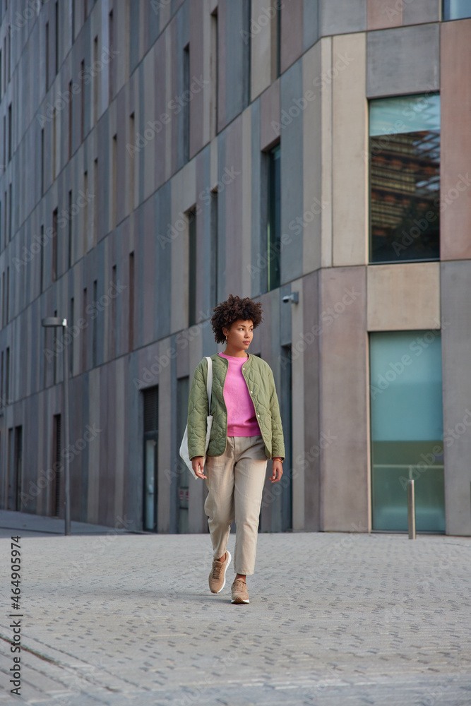 Full length shot of fashionable woman walks outdoors against modern city building carries bag being on her way to necessary place enjoys international vacations explores street architecture.