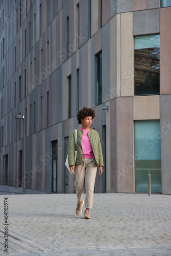 Full length shot of fashionable woman walks outdoors against modern city building carries bag being on her way to necessary place enjoys international vacations explores street architecture. © wayhome.studio 