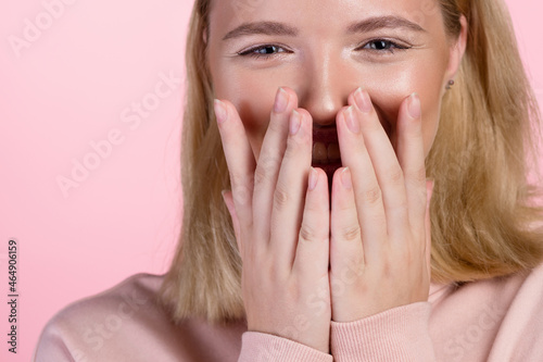 cute young blonde in a pink hoodie laughs merrily covering her mouth and face with her hands. portrait on a pink background, close-up