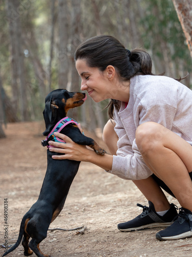 Young caucasian woman hugging and looking with smile to black smooth haired miniature dachshund in forest