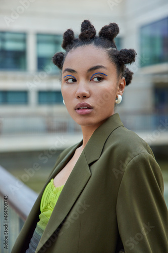 Sideways shot of fashionable girl with hair buns dressed in stylish green jacket enjoys life poses outdoors against blurred background focused into distance. Youth style and appearance concept