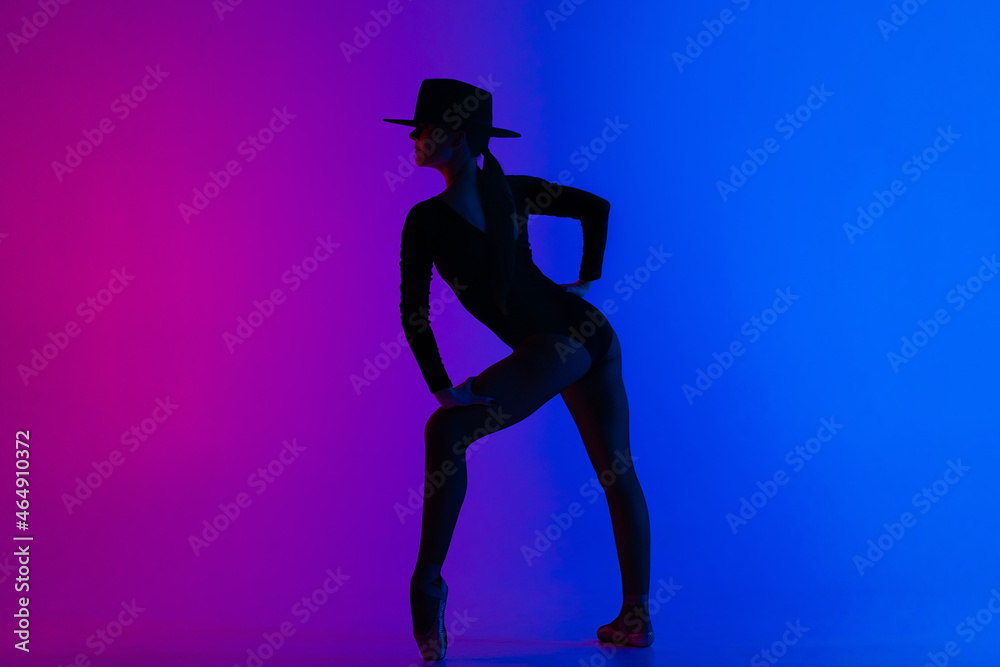 Contemporary ballet dancer woman in hat posing on multi-colored gradient pink blue background. Professional ballerina in sexy costume of femme fatale, solo performance