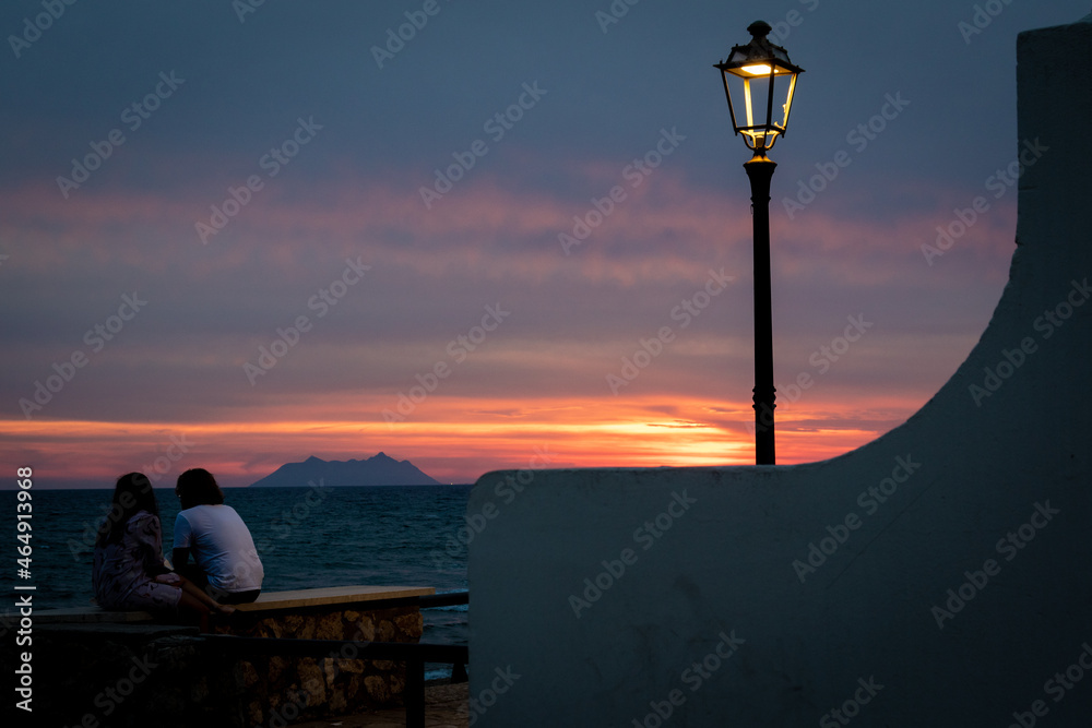 A young loving couple sit in front of a red sunset near the beach