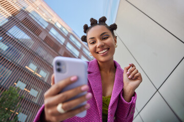Stylish lady with hairstyle dressed in fashionable pink jacket takes selfie on mobile phone for social networks poses against blurred city buildings smiles happily enjoys free time streams video
