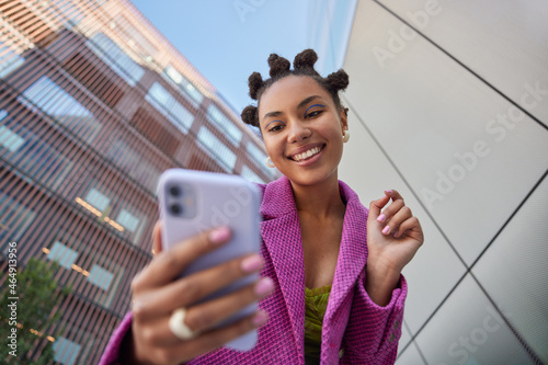 Stylish lady with hairstyle dressed in fashionable pink jacket takes selfie on mobile phone for social networks poses against blurred city buildings smiles happily enjoys free time streams video