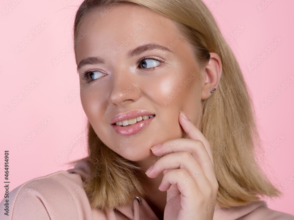 cute young blonde in a pink hoodie smiles happily and touches her face with her hand. cute young blonde in a pink hoodie smiles happily and touches her face with her hand.