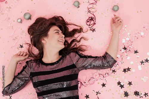 Cheerful girl lies on a pink background among confetti and serpentine, top view.
