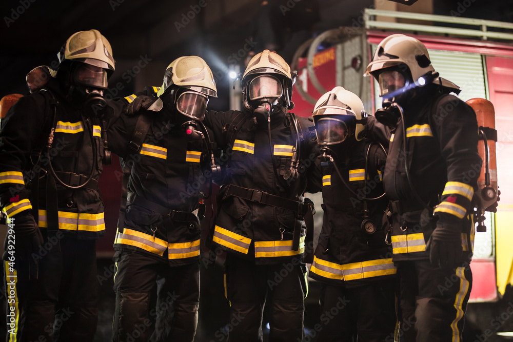 Group of professional firefighters posing. Firemen wearing uniforms, protective helmets and oxygen masks. Smoke and firetrucks in the background. Group of professional firefighters posing. Firemen wearing uniforms, protective helmets and oxygen masks. Smoke and firetrucks in the background.
