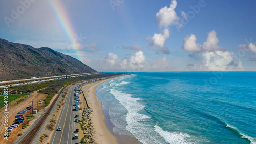  an aerial shot of the vast blue ocean water, lush green farmland, cars on the road, mountain ranges, thick cloud cover with blue sky and a rainbow at Rincon Beach in Ventura County California photo