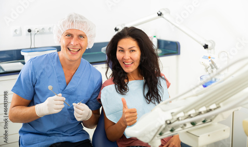 Happy asian woman sitting in dental chair after teeth cure giving thumb up