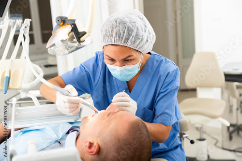 Woman dentist in mask holding dental drill and treating teeth of male patient in dentist office