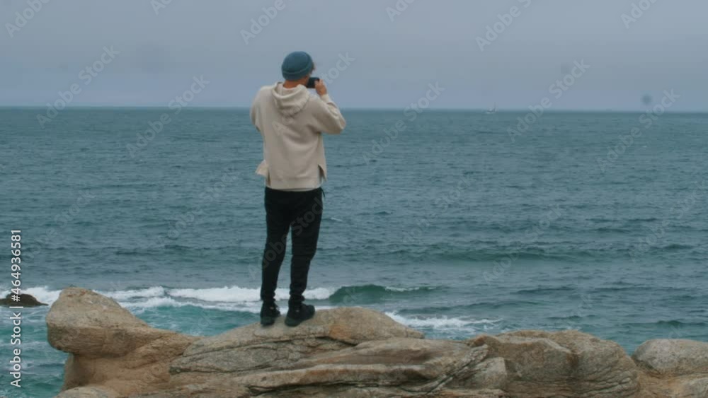 Casual looking Young Man with blue hat taking photo with smartphone in front of ocean. Stand on top of windy cliff overlooking the sea. Human against elements. Tourist enjoy nature. Casual looking Young Man with blue hat taking photo with smartphone in front of ocean. Stand on top of windy cliff overlooking the sea. Human against elements. Tourist enjoy nature.