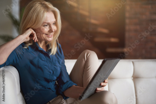 Smiling mature woman watching video using digital tablet pc, relaxing on a couch at home in the living room