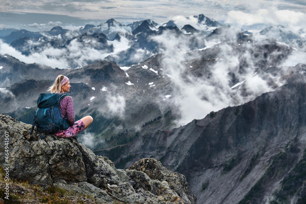 Adventurous woman sitting on cliff over mountains and clouds relaxing and meditating. Whistler. Garibaldi Provincial Park. British Columbia. Canada