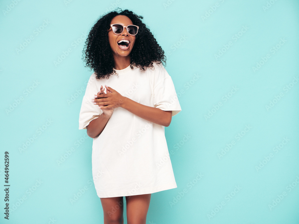 Beautiful black woman with afro  hairstyle. Smiling model in long t-shirt clothes. Sexy carefree female posing near blue wall in studio. Tanned and cheerful in sunglasses - obrazy, fototapety, plakaty 