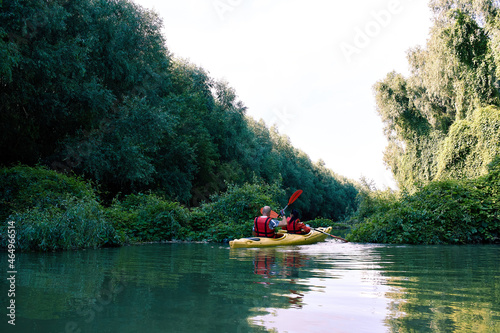 Family couple man and a woman is kayaking in yellow kayak on a river near big green trees and wild grapes. Back view on couple kayaking on a river at summer