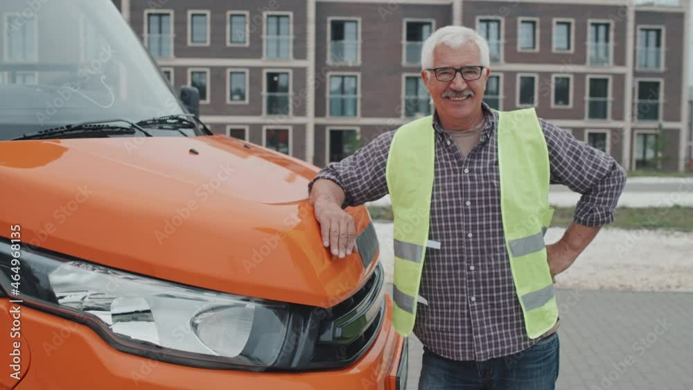 Medium handheld portrait of friendly senior bus driver in acid green vest smiling to camera standing outdoors leaning on orange bonnet of modern passenger bus