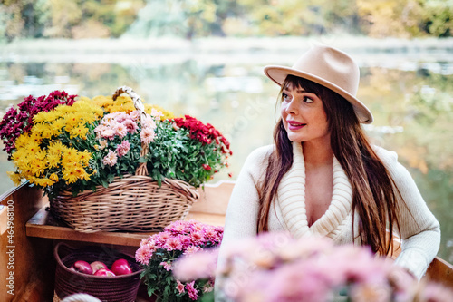 young beautiful plus size model in autumn brown warm coat and hat sitting in boat on pond decorate with chrysanthemums and apples in autumn park, concept of season leaf fall, romance and unusual date © klavdiyav