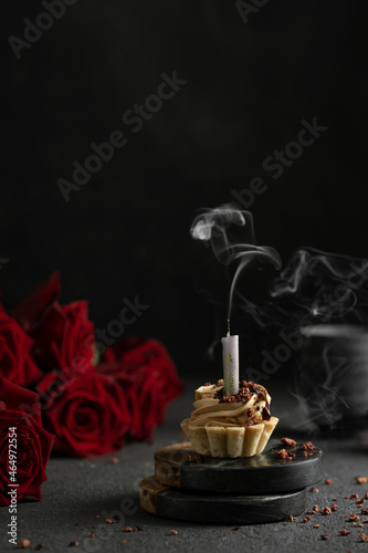 Little cake with candle and smoke with red roses on black table. Mystic dramatic birthday concept. Dark mood greeting birthday card.