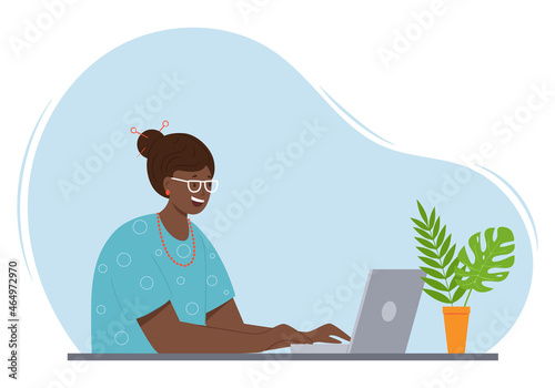 A happy adult woman with dark skin and hair is sitting at a table with a laptop. An adult modern lady in glasses and beads communicates at the computer. Color vector illustration on a spot background.