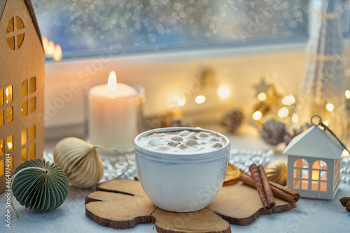 Chocolate drink with marshmallow, cinnamon and milk froth. Christmas seasonal beverage on decorated windowsill. Wintertime. Candles and garland blurred background, selective focus