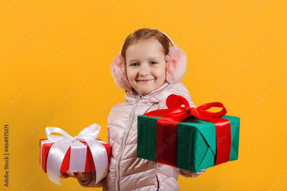 Happy little girl holds out a box with a gift. Child in a jacket and warm headphones on a yellow isolated background. Christmas new year winter concept