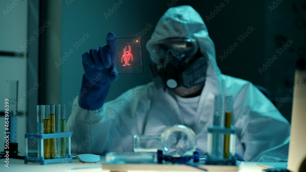 Scientist in ppe suit looking at small transparent tablet with biohazard hologram in lab Scientist in ppe suit looking at small transparent tablet with biohazard hologram in lab