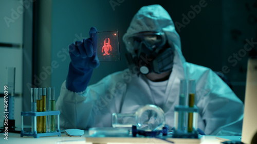 Scientist in ppe suit looking at small transparent tablet with biohazard hologram in lab