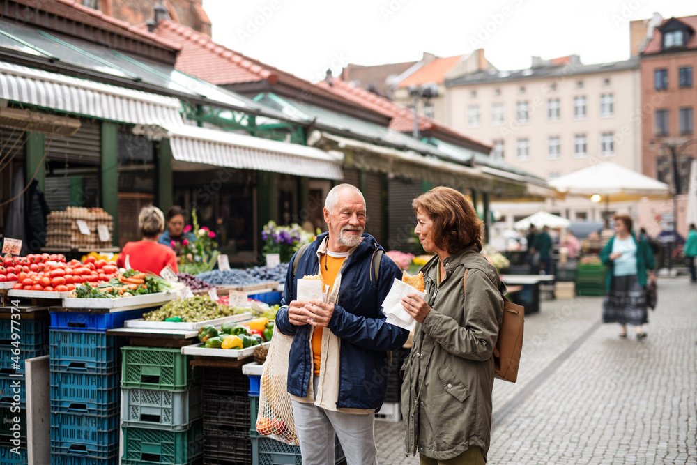 Portrait of happy senior couple tourists with snack in town on outdoor market.
