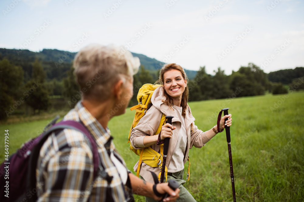 Happy mid adult woman with trekking poles hiking with active senior mother outdoors in nature.