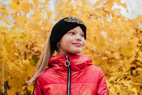 Smiling little girl standing near yellow leaf bush and hiding. Happy child playing in autumn forest. Smiling kid having fun outdoors. Red jacket  knitted headband  black pants.