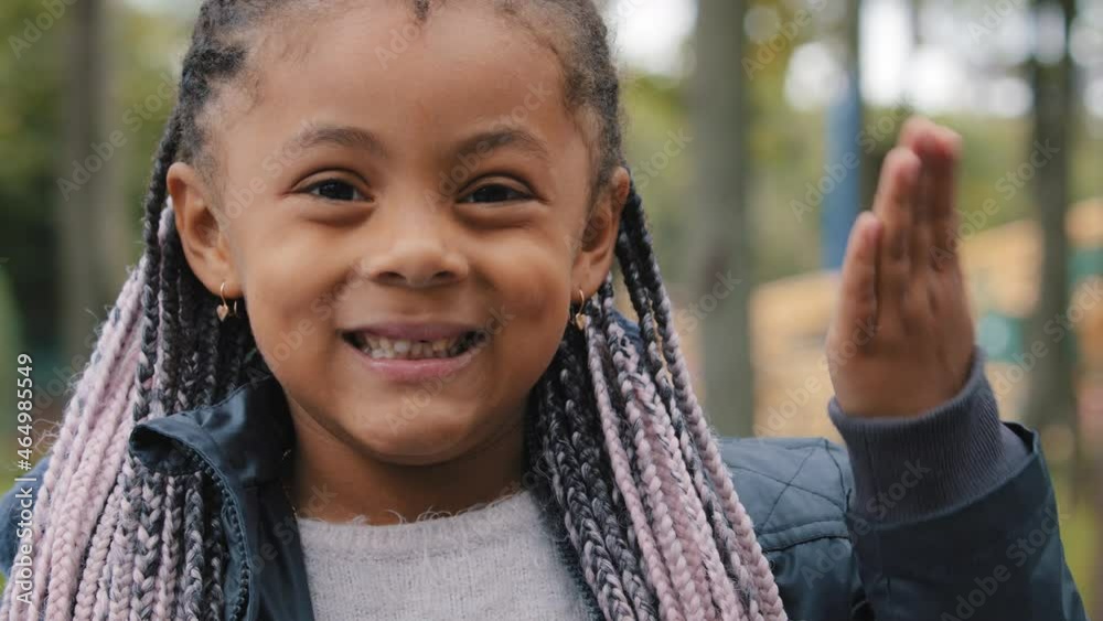 Little cute girl afro american mixed race child kid baby daughter with hairstyle braids looking at camera smiling wide toothy waving hello greeting video call web camera view portrait outdoors in city Little cute girl afro american mixed race child kid baby daughter with hairstyle braids looking at camera smiling wide toothy waving hello greeting video call web camera view portrait outdoors in city