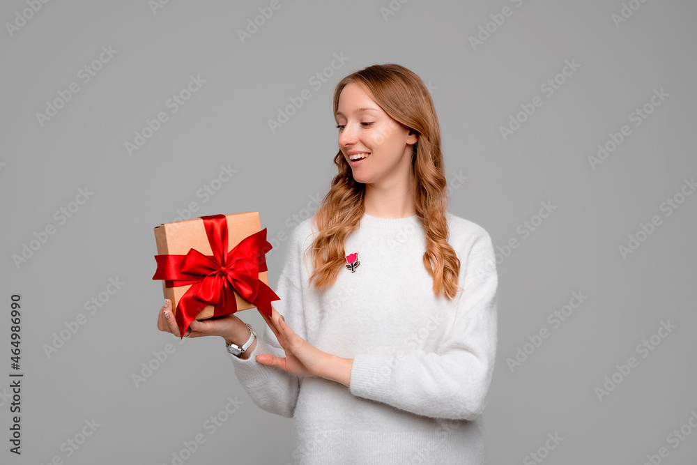 Young happy woman with blonde curly hair posing on gray background with gift box decorated with red ribbon. New Year Women's Day birthday holiday concept