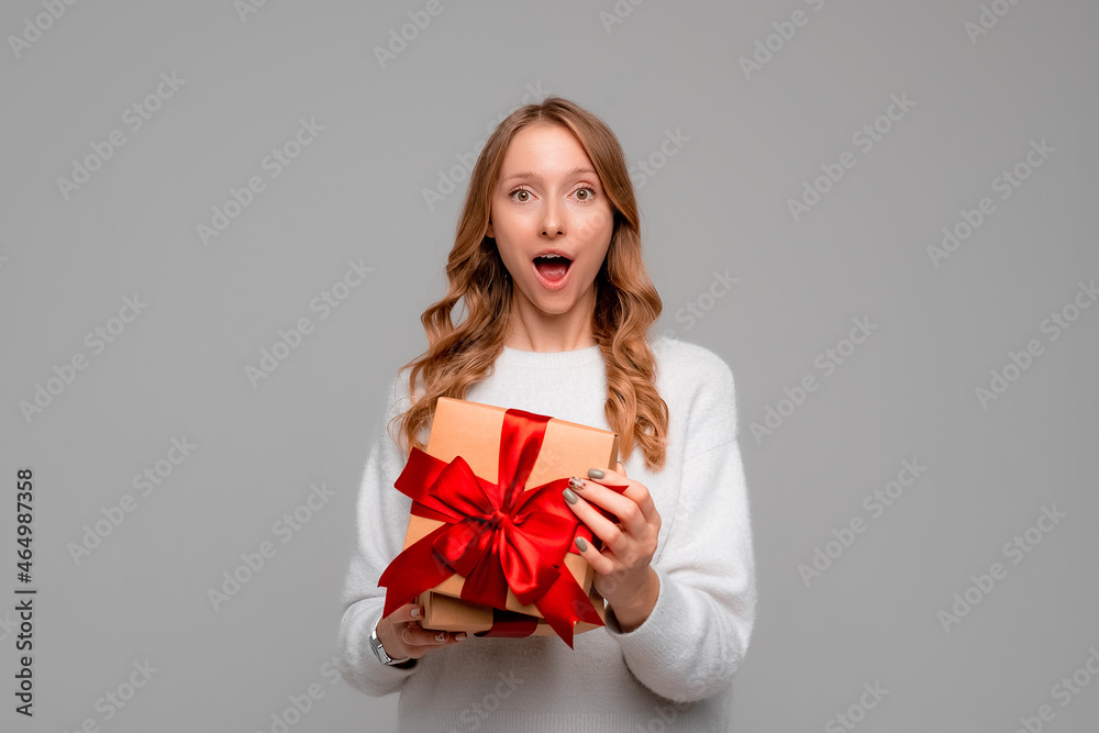 Beautiful young woman holding in hands unboxed present and looking at camera with surprised expression, standing over gray background. New Year Women's Day birthday holiday concept Beautiful young woman holding in hands unboxed present and looking at camera with surprised expression, standing over gray background. New Year Women's Day birthday holiday concept