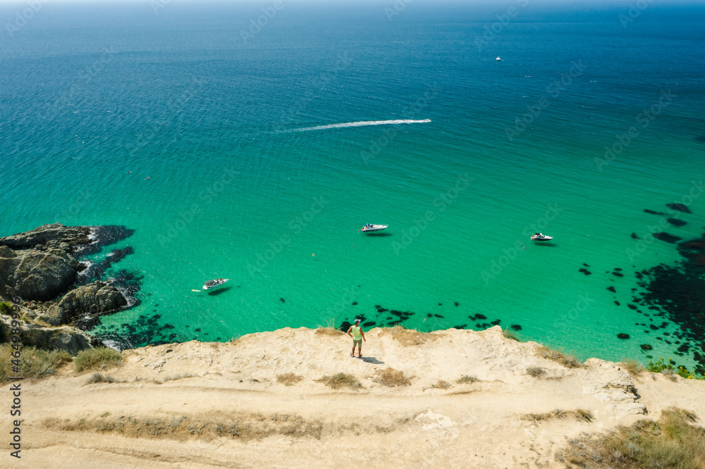Obraz premium Man traveler on the edge in front of Bounty beach with crystal clear azure sea on a sunny day. Cape Fiolent in Sevastopol. Aerial view