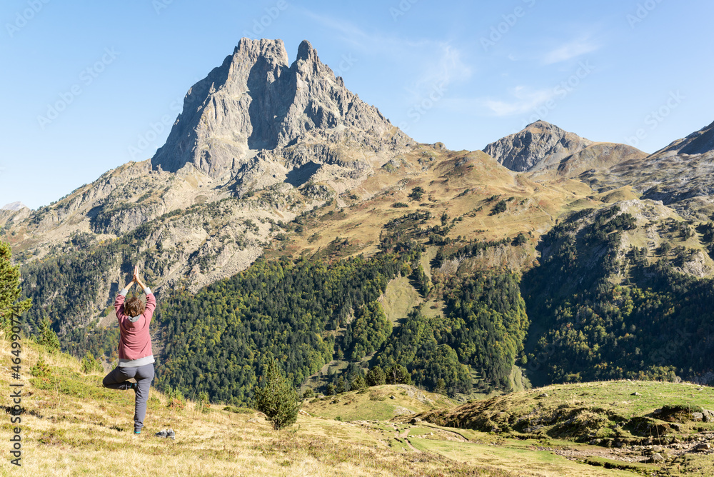 Back view woman practising yoga on a green meadow with Pic du Midi d'Ossau in front of her in Pyrenees. Yoga at the mountains and harmony with nature. Back view woman practising yoga on a green meadow with Pic du Midi d'Ossau in front of her in Pyrenees. Yoga at the mountains and harmony with nature.