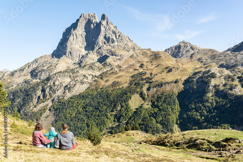 Family trekking day at french Pyrenees. Father, mother and baby resting on a meadow while are looking at Pic du Midi d'Ossau. Mountain travel vacation.