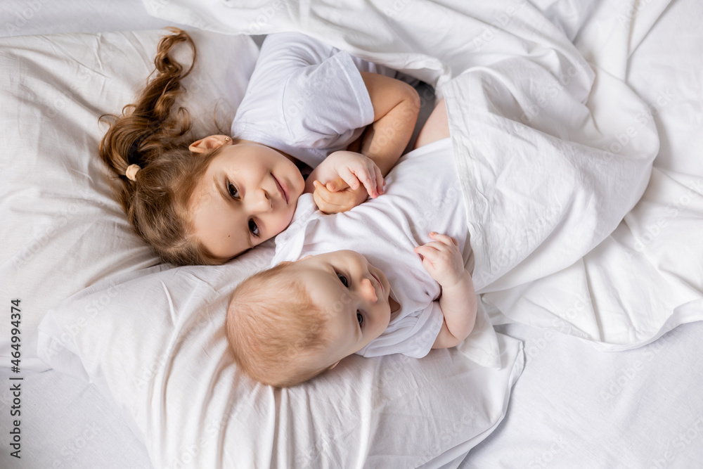 little girl and baby in white clothes are lying in bed on white bed linen. brother and sister bask in bed. children's sleep and rest. hygge, lifestyle. space for text. High quality photo little girl and baby in white clothes are lying in bed on white bed linen. brother and sister bask in bed. children's sleep and rest. hygge, lifestyle. space for text. High quality photo