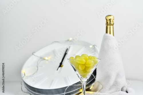 Glass with grapes on the background of a blurry clock and a bottle of champagne. Spanish tradition of eating twelve grapes to celebrate New Year.