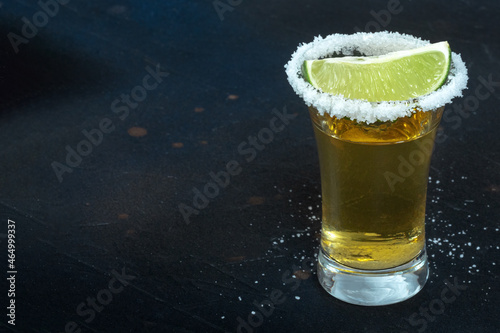 Golden tequila shot, with a salted rim and a lime slice, on a black background with copy space