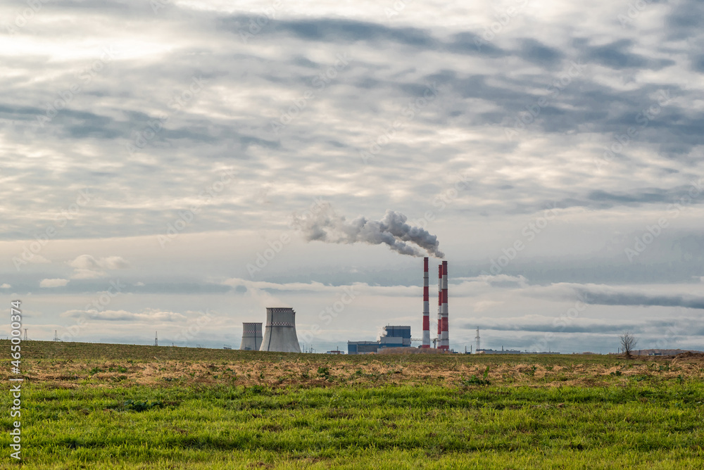 Green field against the background of clouds and blue sky, behind it a thermal power plant with smoking chimneys