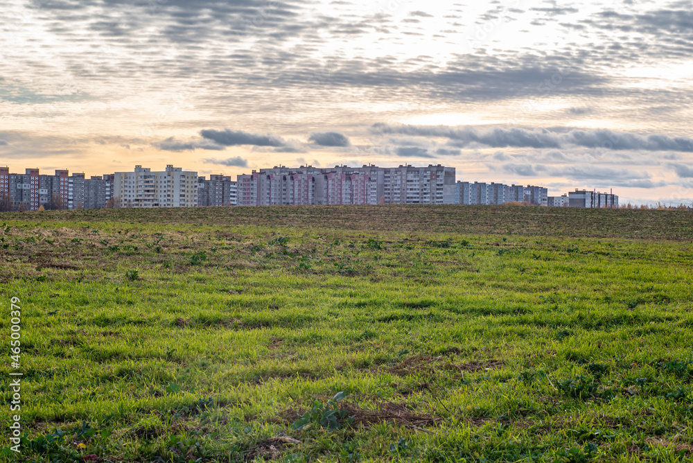 Green field and sunny evening sky with clouds, background of concrete multi-storey houses