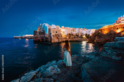Young woman on the rocks in front of the Apulian village of Polignano a Mare at sunset (blue hour)
