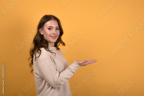 A young and attractive Caucasian brunette girl with wavy hair holding a empty space in her palm on an orange studio background. © Павел Костенко