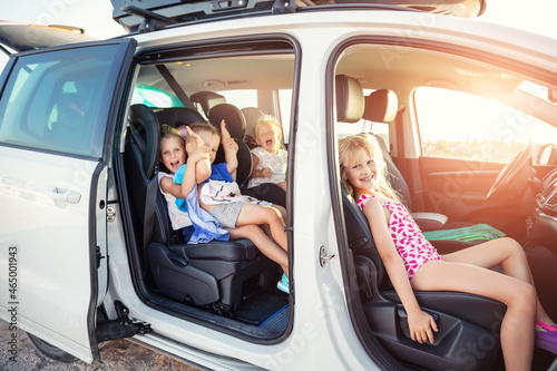 Group of four cute adorable little happy caucasian children enjoy having fun sit in minivan going to sea beach road trip on hot summer day. Kids in swimsuit in car with side open door against sun photo