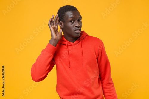 Curious young african american man guy in red streetwear hoodie posing isolated on yellow wall background in studio. People lifestyle concept. Mock up copy space. Try to hear you with hand near ear.
