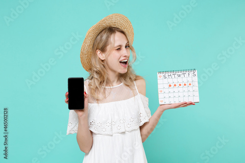 Blinking girl in hat hold periods calendar for checking menstruation days isolated on blue turquoise background. Medical healthcare gynecological concept. Hold mobile phone with blank empty screen.