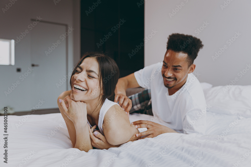 Fototapeta premium Laughing young couple two family in casual white clothes lying in bed on stomach man tickling woman fooling around time together in bedroom lounge home house wake up dream be lost in reverie good day.