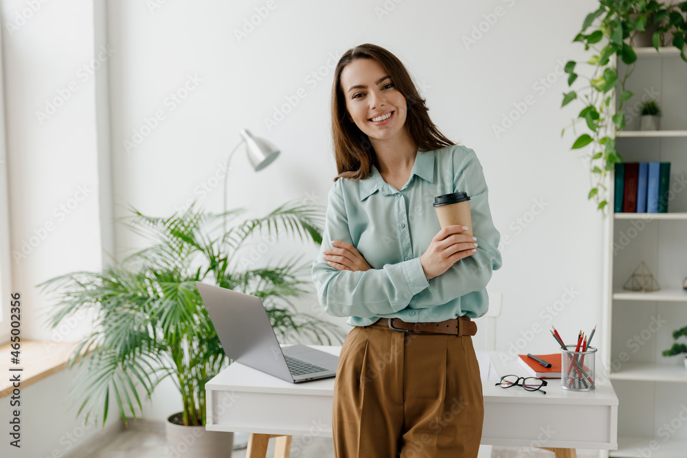 Young successful employee business woman 20s in blue shirt work hold takeaway delivery craft paper cup coffee to go stand at desk with laptop pc computer at office indoors. Achievement career concept. Young successful employee business woman 20s in blue shirt work hold takeaway delivery craft paper cup coffee to go stand at desk with laptop pc computer at office indoors. Achievement career concept.